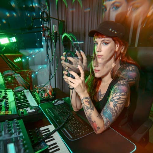 Sishiva in the studio, holding headphones beside a synthesizer